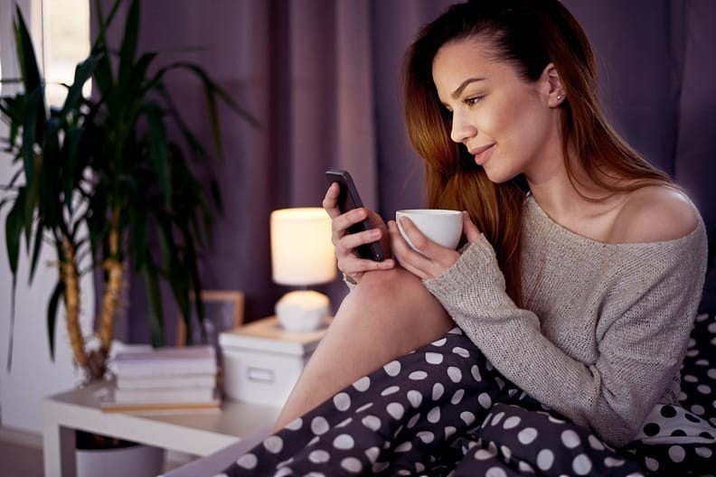 A cheerful woman sitting in a bedroom while holding her phone, smiling as she drafts a text message to her partner