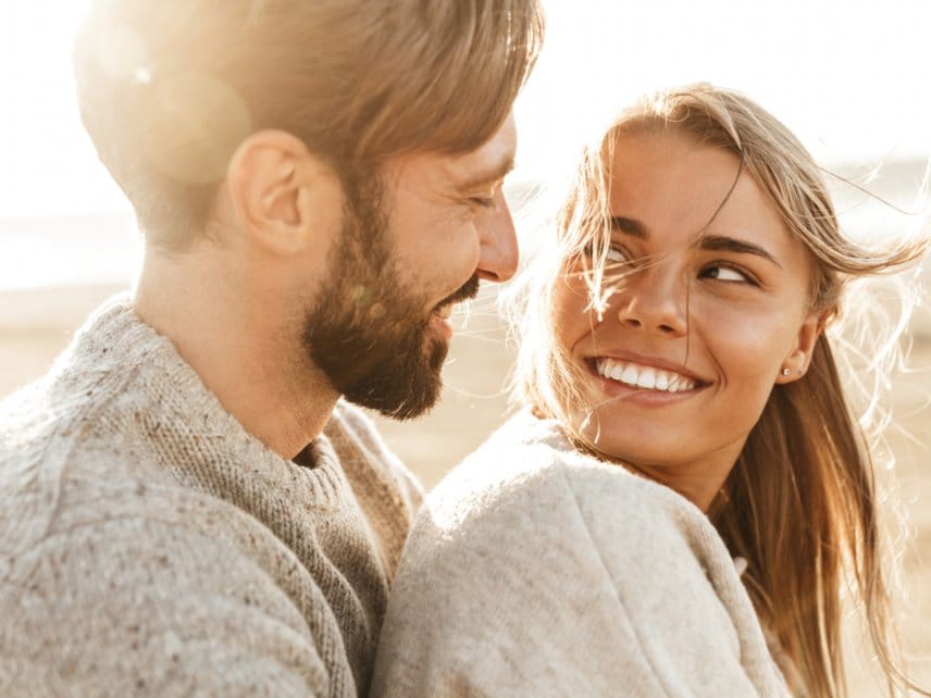 A bubbly woman and man, smiling at each other and standing on a beach.