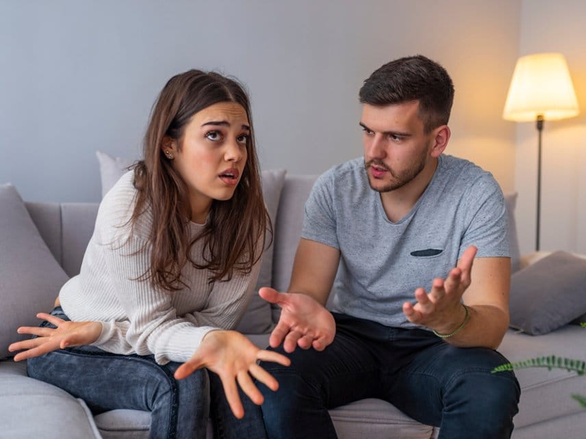 A girlfriend and boyfriend sitting together on a living room couch, having a serious conversation about communication issues and emotional tension in their relationship